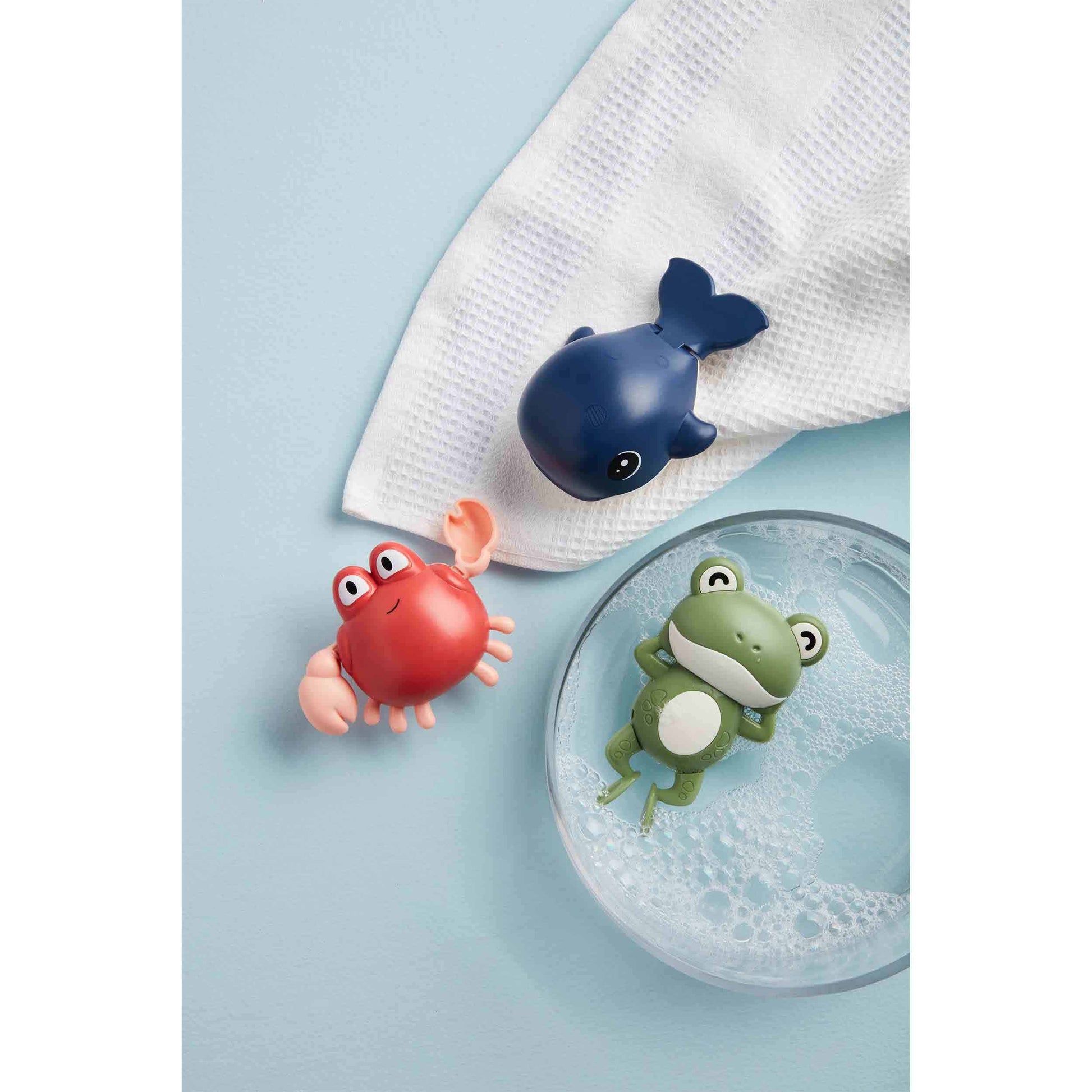 3 wind-up toys on a blue background, frog is in a dish of water, crab and whale are on a white towel.