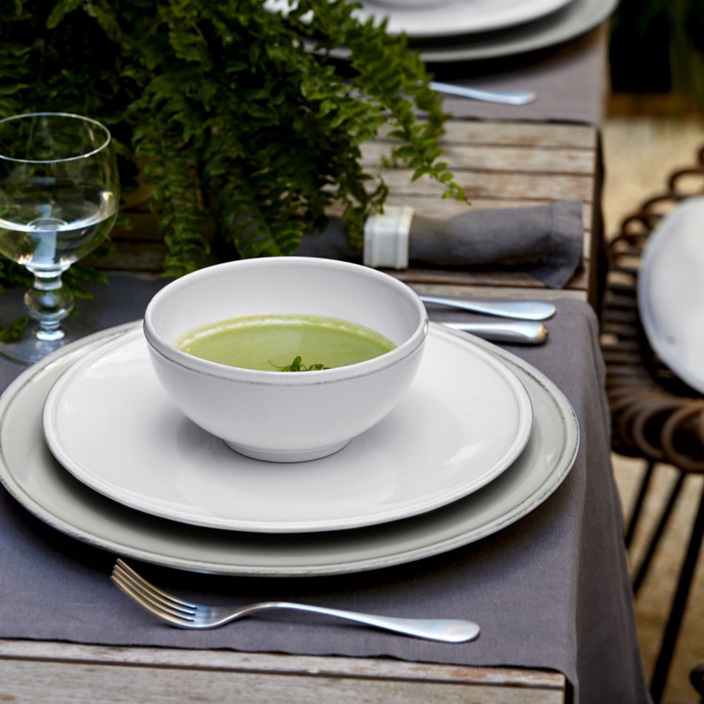 side view of table setting with white bowl on top filled with soup, silverware, glasses, and a fern.