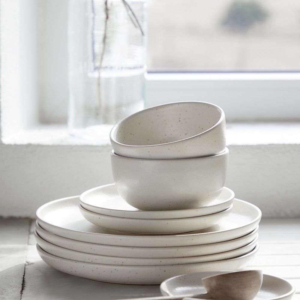 stacked vanilla dinnerware displayed in front of a window next to a vase with dried flowers on a white countertop
