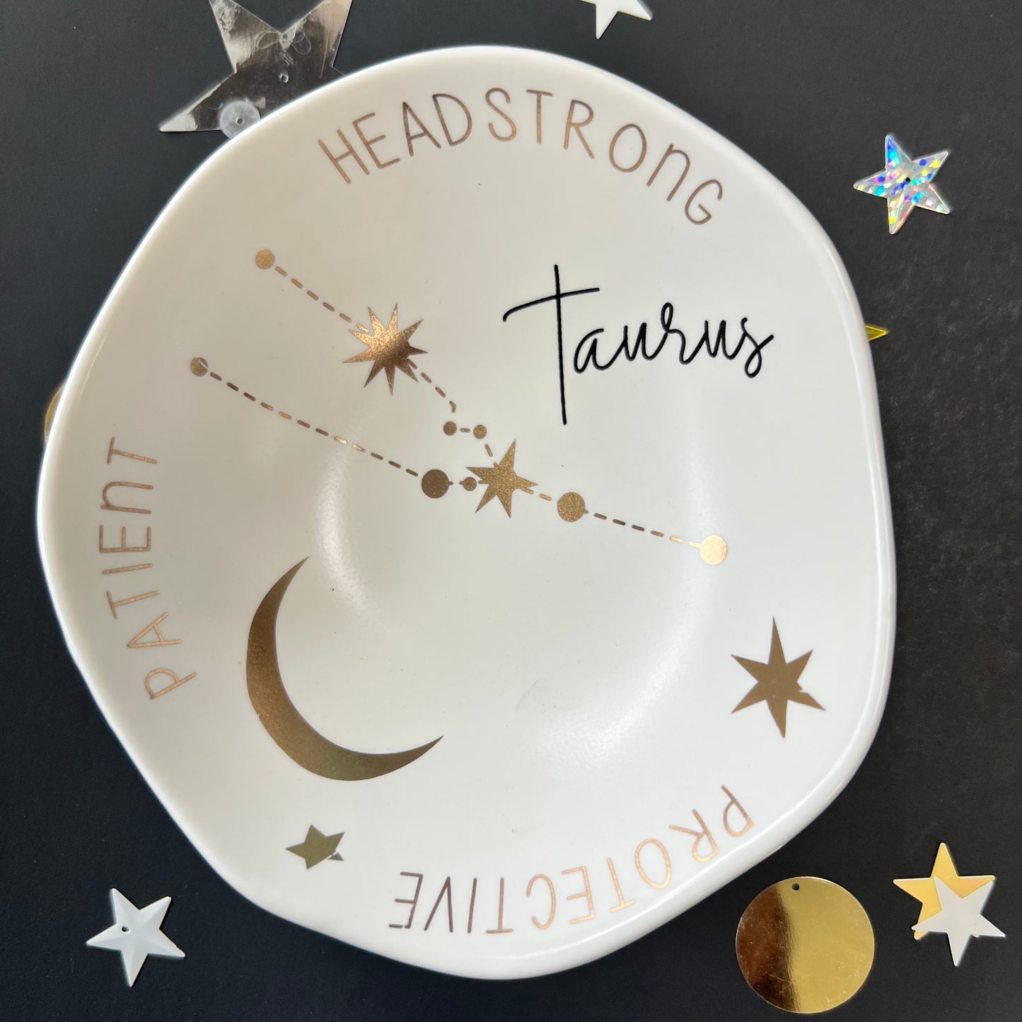 cream dish with gold stars and "Taurus Headstrong Protective Patient" around the inner rim on a black background with scattered stars and orbs.