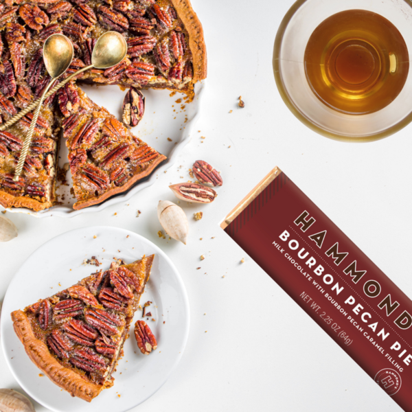 the bourbon pecan pie milk chocolate candy bar displayed next to a piece of pecan pie a cut pecan pie and a glass of bourbon on a white background