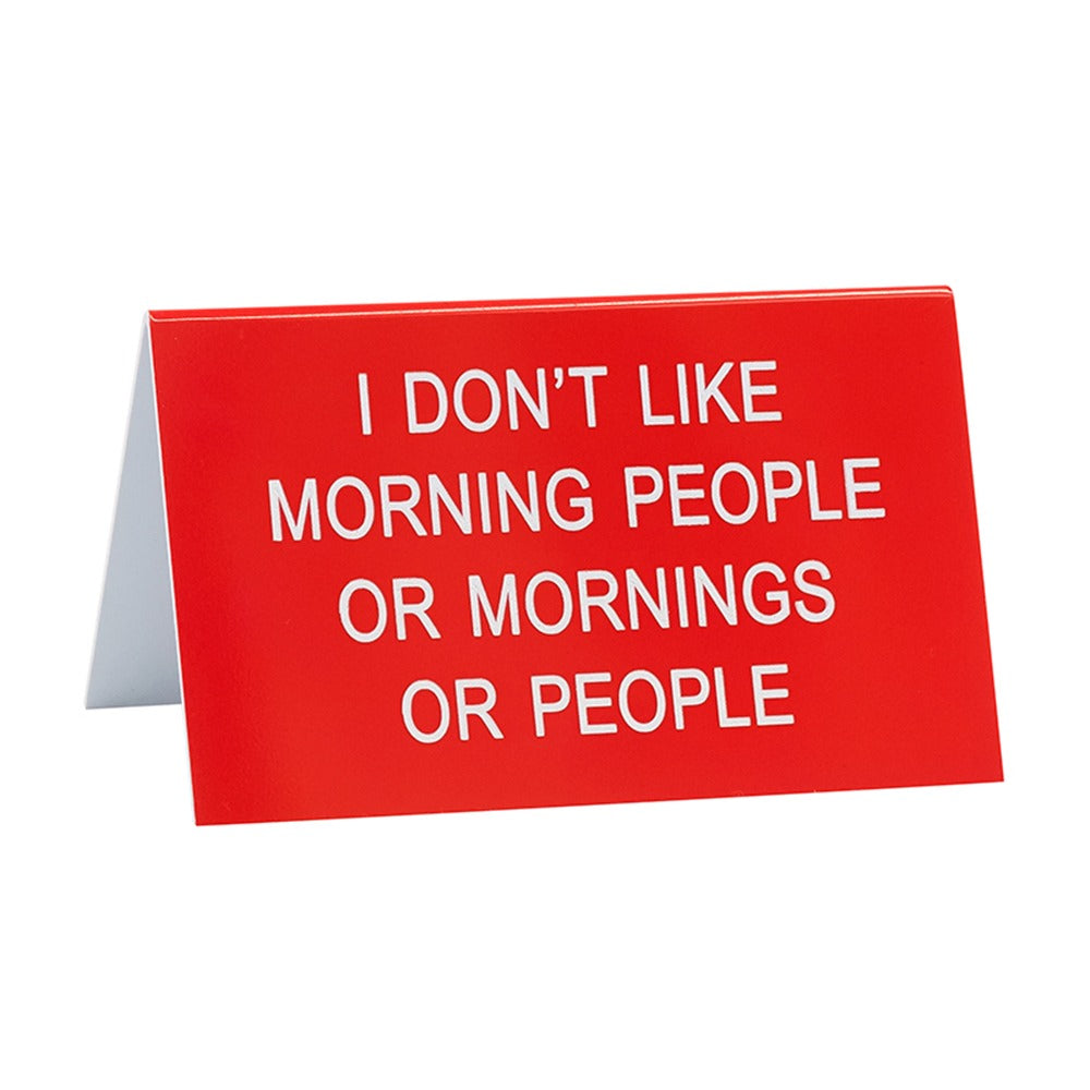 red funny desk sign with text "I don't like morning people or mornings or people" against a white background