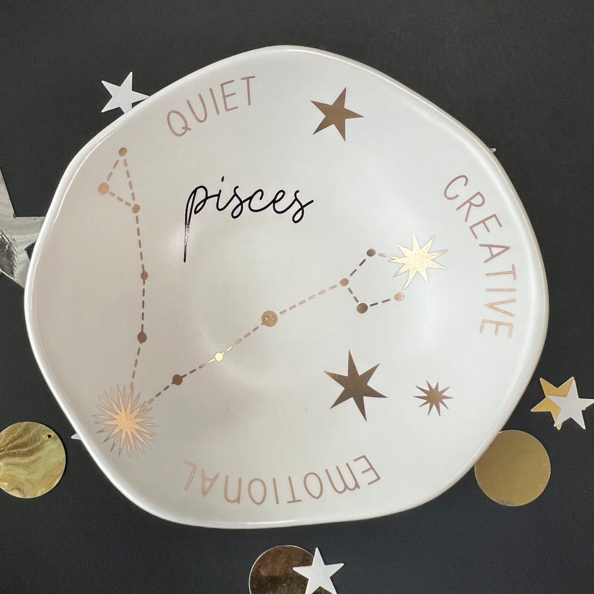 cream dish with gold stars and "Pisces Quiet Creative Emotional" around the inner rim on a black background with scattered stars and orbs.