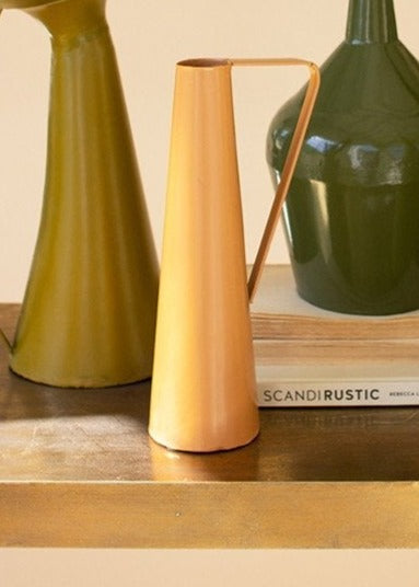 peach metal vase with handles displayed on a table next to two vases stacked books against a pale pink background