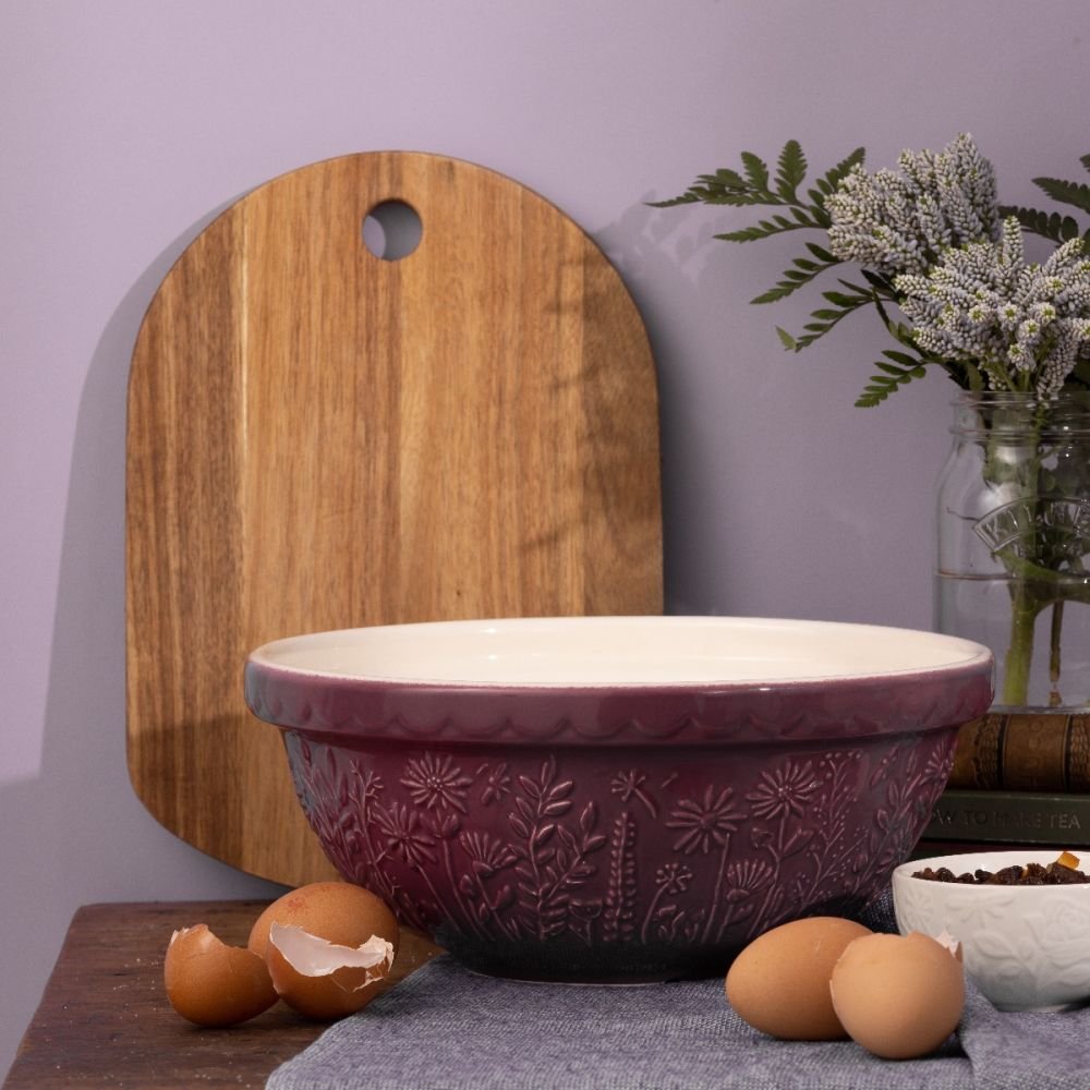 purple bowl on a countertop with wood board, eggs, and flowers.