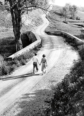 front of card is a photograph of a man and woman holding hands and walking down a dirt road over a bridge