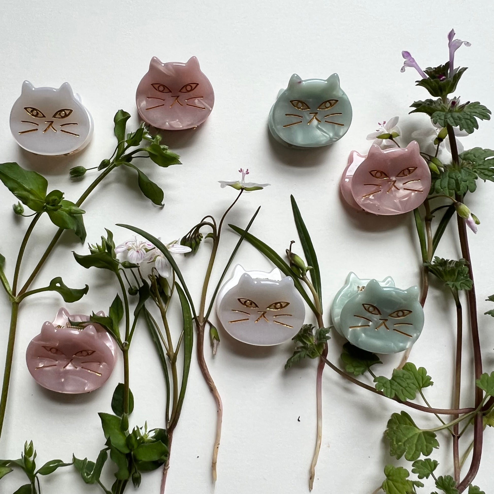 3 colors of cat face hair clips arranged on a white background with greenery and flowers.