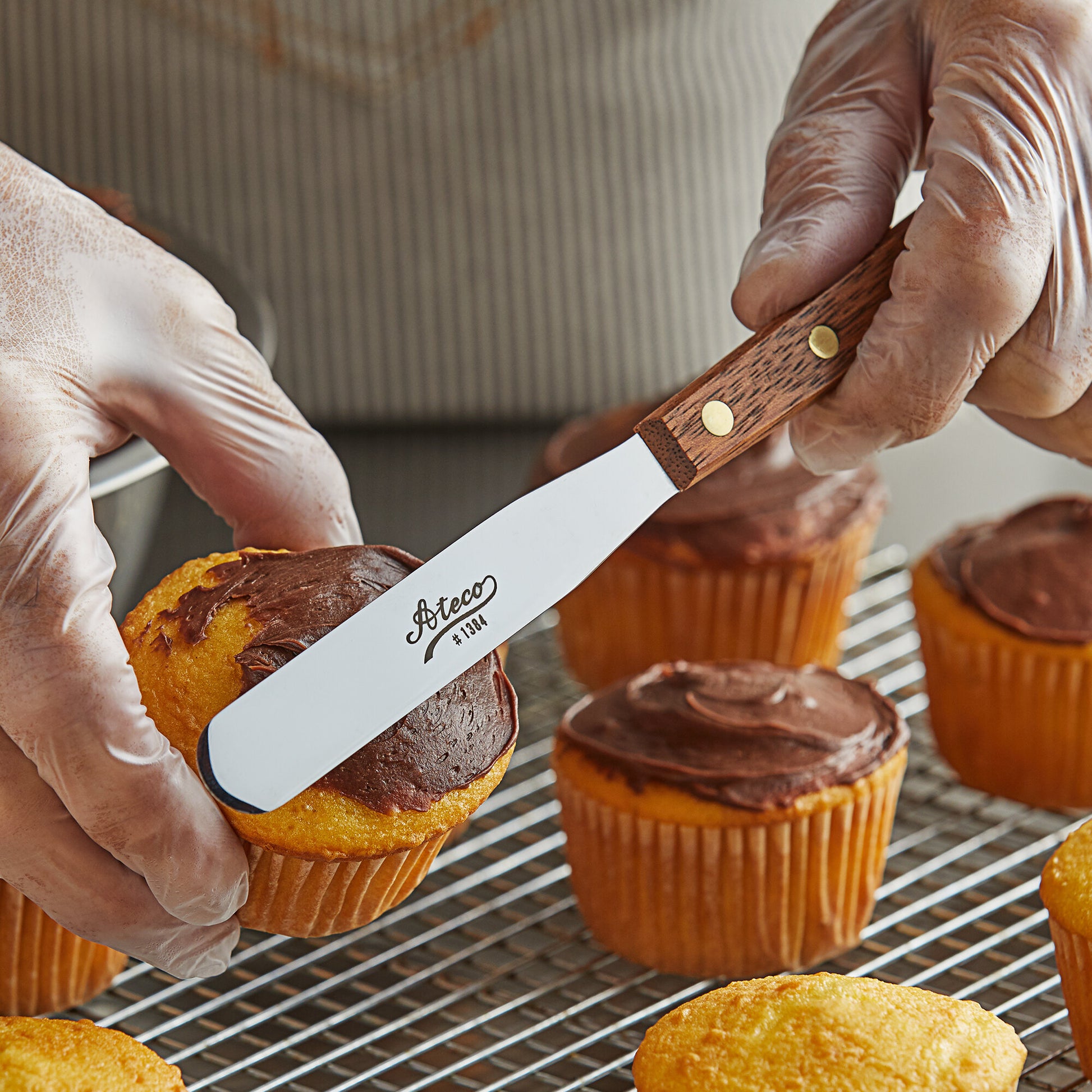 illustration of the wooden handle icing spatula icing a batch of cupcakes with a person in the background