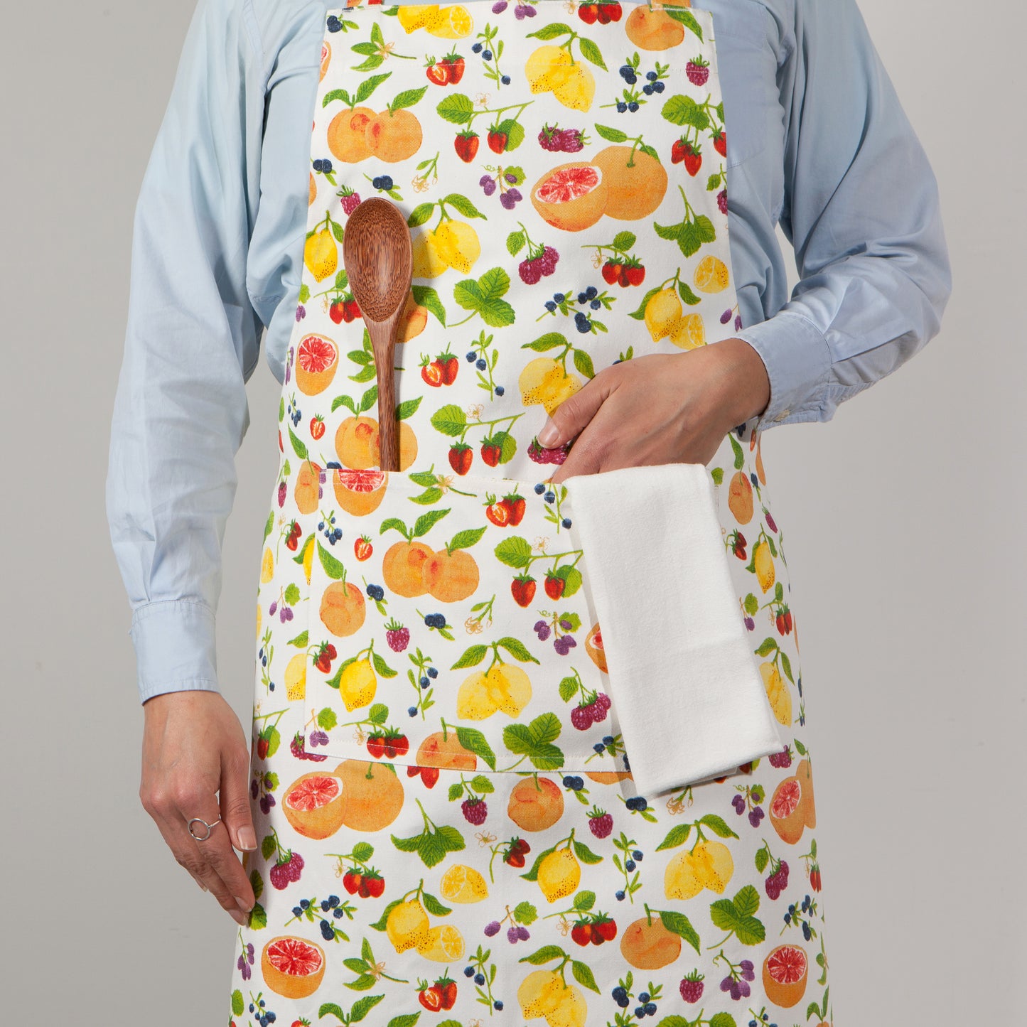 person wearing white apron printed with all-over fruit pattern. person has hand in pocket with dishtowel draped in it and a wooden spoon in the other pocket.