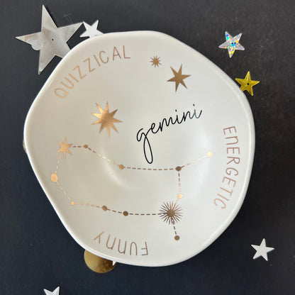 cream dish with gold stars and "Gemini Quizzical Energetic Funny" around the inner rim on a black background with scattered stars and orbs.