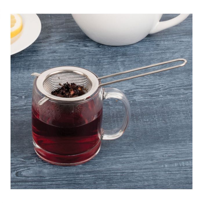 double ear conical tea strainer filled with tea leaves resting on a glass mug filled with tea on a gray wooden surface