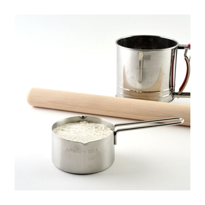 Stainless Steel Measuring Cup filled with flour and a rolling pin and sifter next to it.
