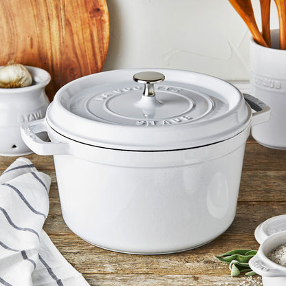 white dutch oven on wooden countertop with kitchen tools in the back ground.
