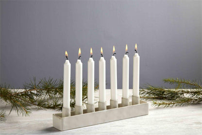 ceramic taper older with 7 lit candles set on a table with sprigs of greenery.