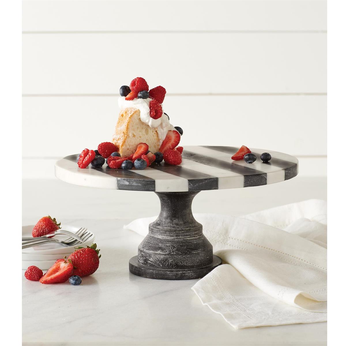 black and white stripped marble cake pedestal displayed with blueberries, strawberries, and slice of cake next to stacked plates and white napkins against a white backsplash in a kitchen