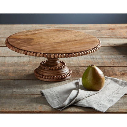 beaded wooden cake stand displayed next to a towel and mango on a rustic wood slat table