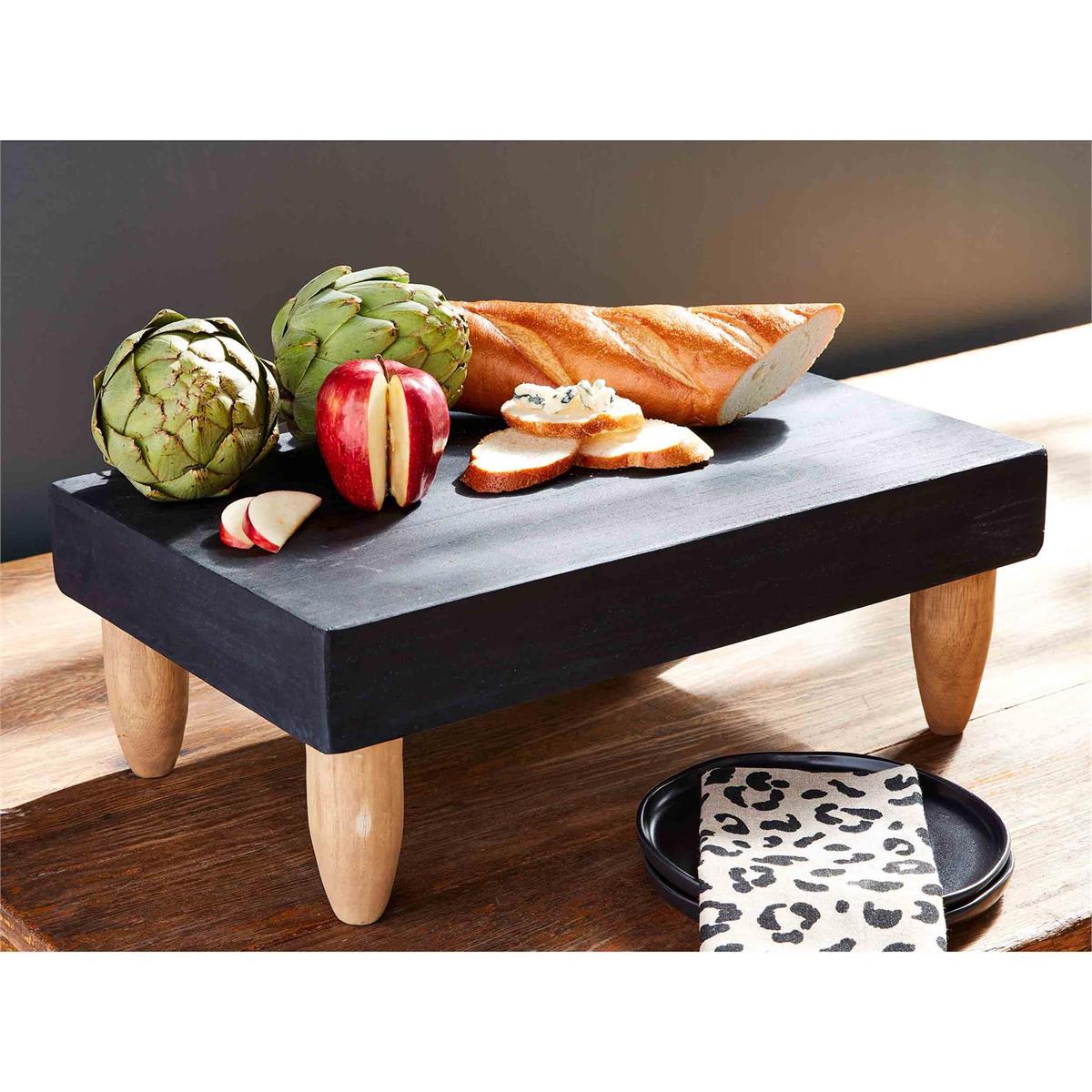 black footed serving board displayed with a beget, artichokes, and apple next to a stack of black plates and cheetah print towel on a wooden surface