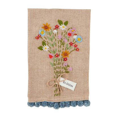 bloom floral embroidered towel is tan with a blue pom trim against a white background