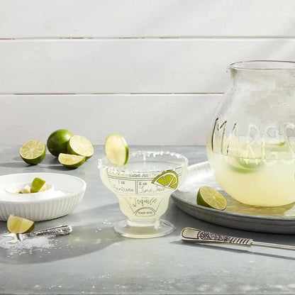 margarita recipe glass displayed on a gray surface next to a margarita pitcher, tin round tray, salt rimmer, and limes