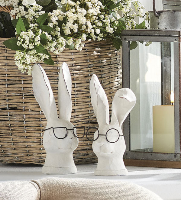 2 white bunnies wearing black glasses displayed in front of a basket of flowers and a lantern with a lit candle in it.