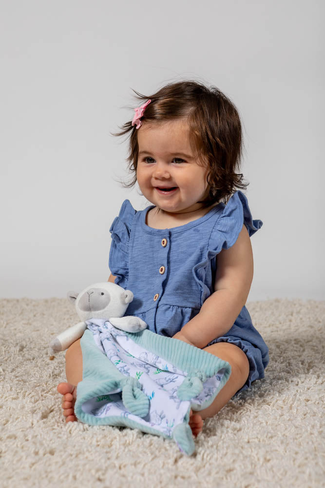 a little girl wearing a blue dress holding the little knotties lamb blanket while sitting on a cream rug