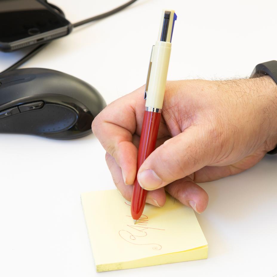 a person writing with the red vintage multi pen on a yellow sticky notes next to a computer mouse on a white surface