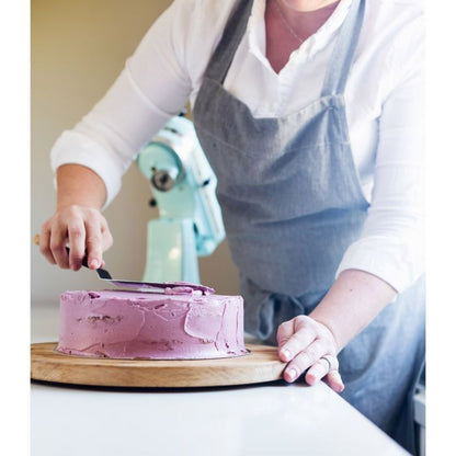 a person decorating a cake with the offset icing spatula on a wooden cake plate with a mixer in the background