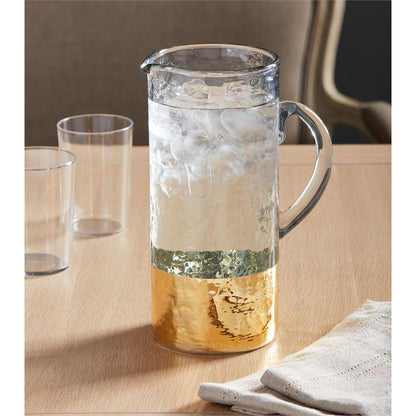 gold hammered glass pitcher displayed next to two glass and folded napkins on a light wood table