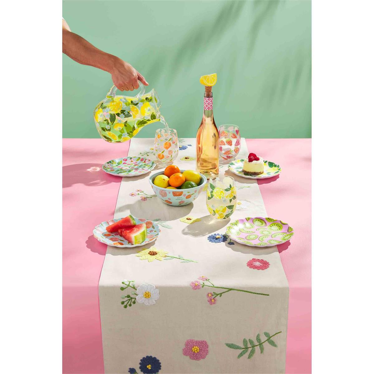 a person using the lemon pitcher to fill up glasses that are sitting in a table setting next to plates, bowl, and wine bottle on a pink table cloth with a floral runner on top