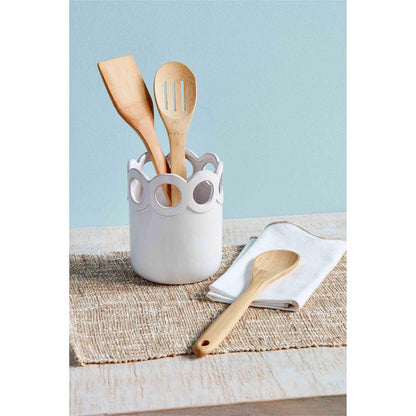 scalloped utensil holder displayed with three wooden utensils on a tan table runner next to a white towel against a pale blue background