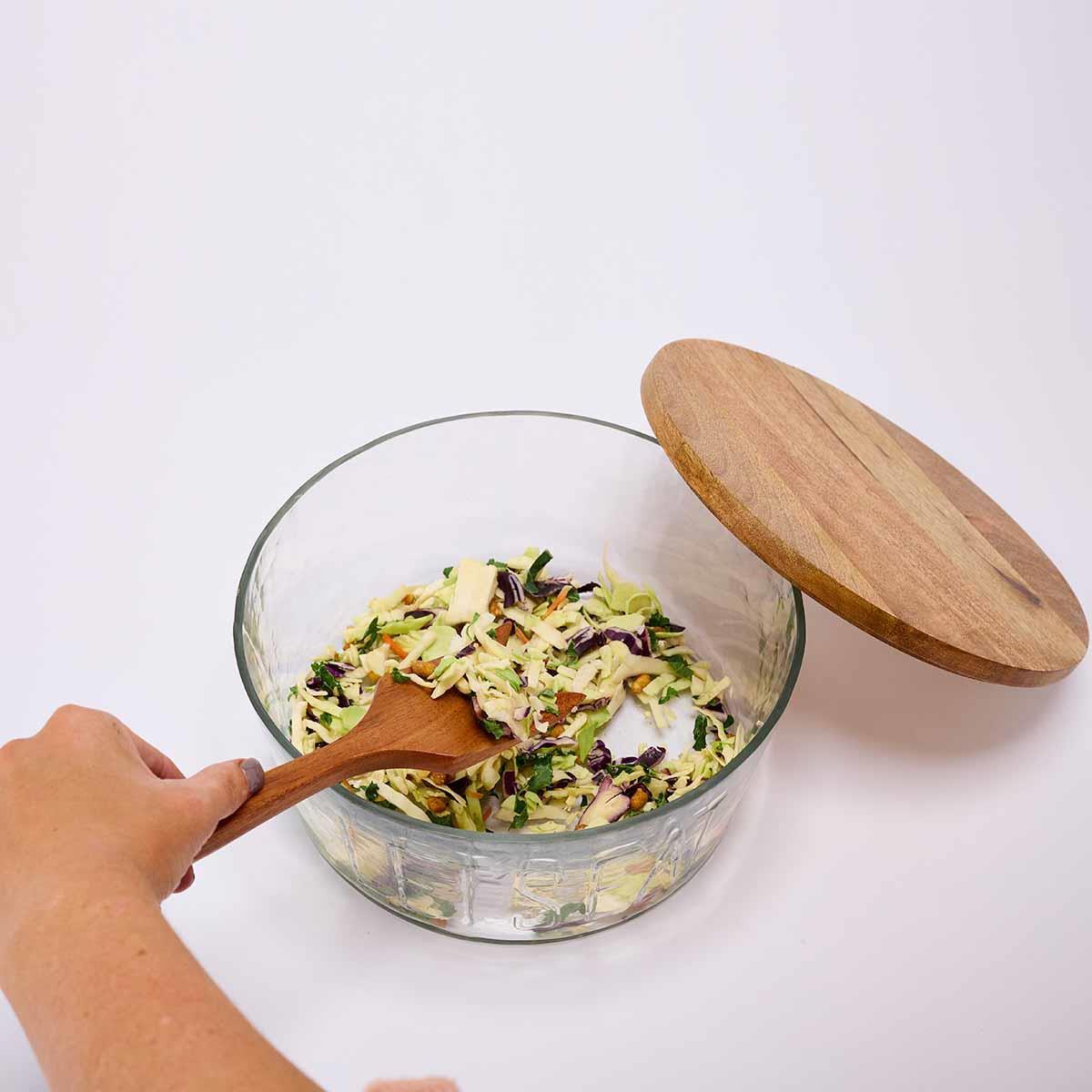 a persons hand stirring a salad in the let's eat glass bowl set on a white background