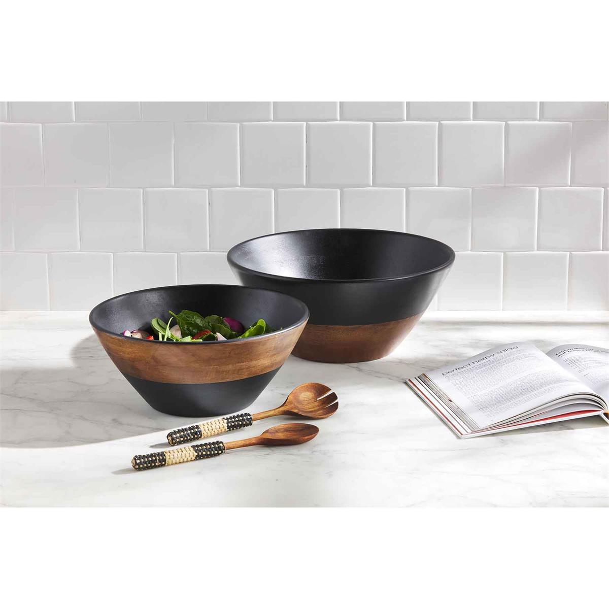 small and large two tone wood bowls filled with salad and displayed next to salad servers and a cookbook on a white marble surface