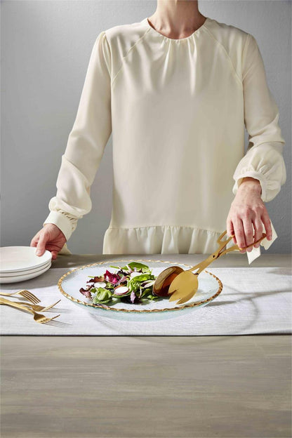a person illustrating the use of the tongs with salad in the gold edge serving bowl displayed next to a stack of salad plates and forks