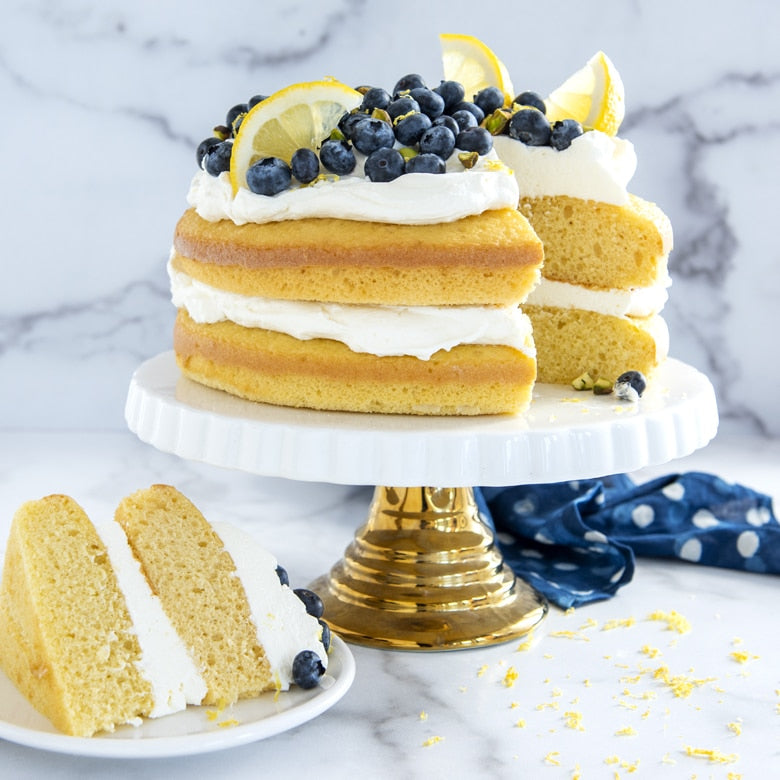 two layer cake on pedestal topped with cream, blueberries, and lemons.