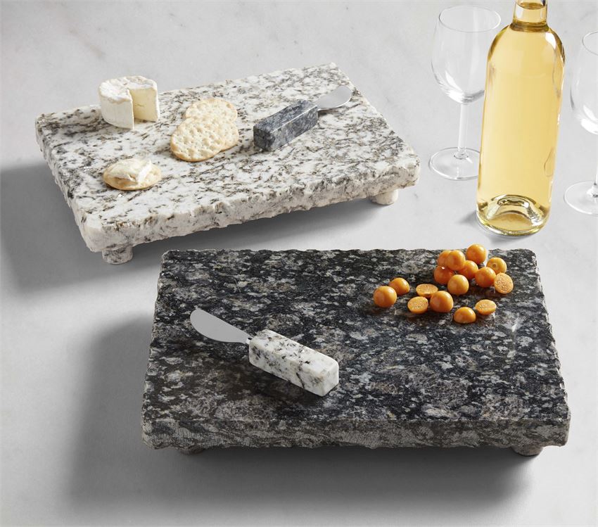 grey granite board with cheese and crackers o it and black granite with fruit on it arranged on a table with wine bottle and glasses.