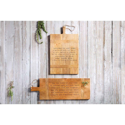 both styles of mango wood board with handles displayed on a whitewashed wood slat surface next to sprigs of greenery