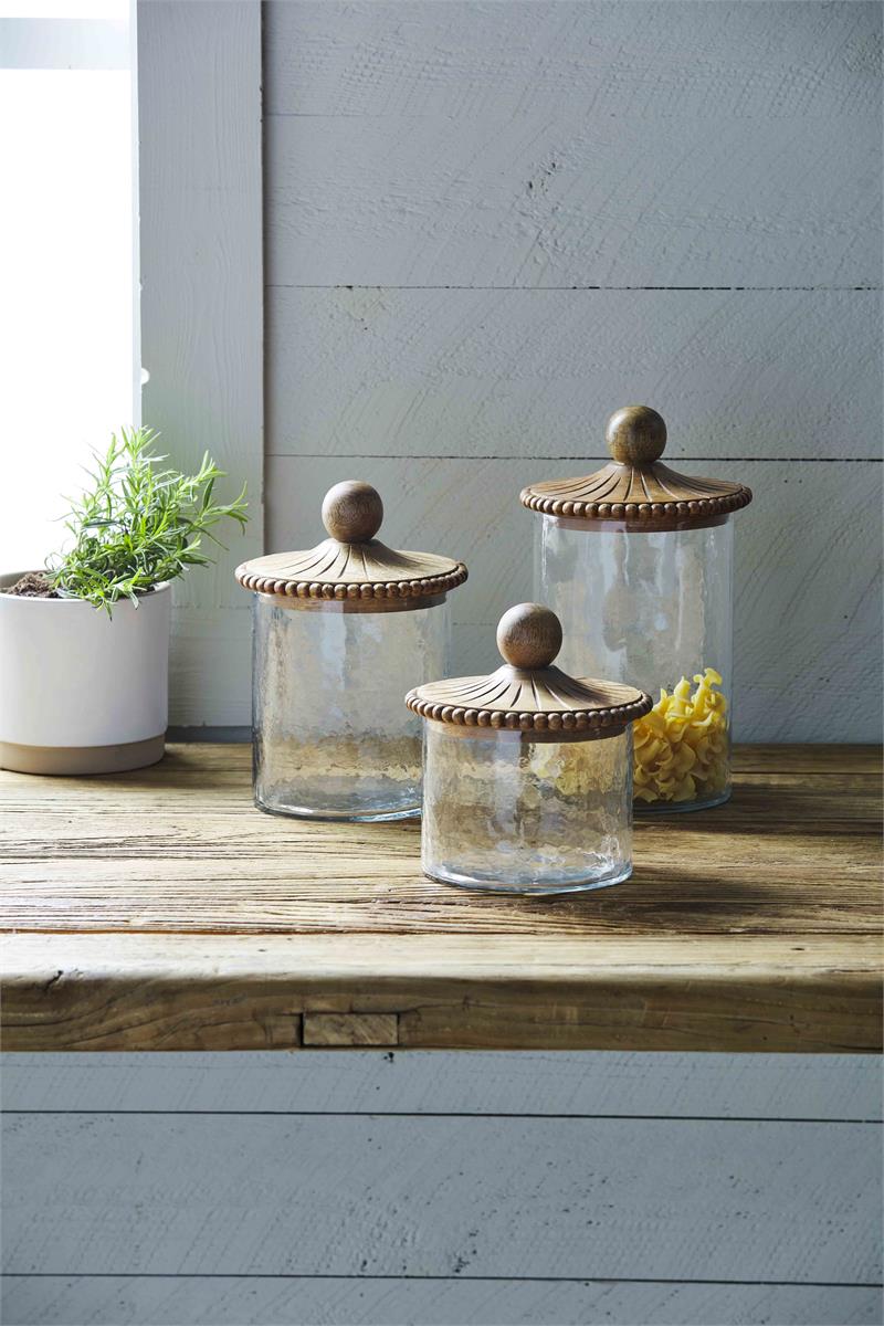 3 sizes of glass canisters, largest one has pasta in it, on a wooden counter near a window with a plant.