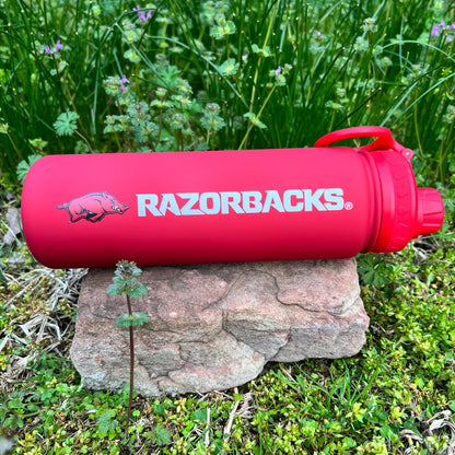red water bottle with razorback logo and text "razorback" in white lettering on a rock with greenery in the background.