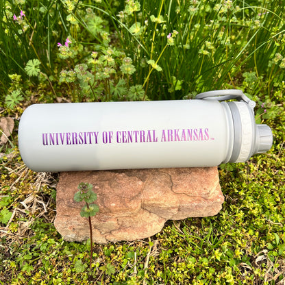 grey water bottle with "university of central arkansas" on purple lettering on a rock with greenery in the background.