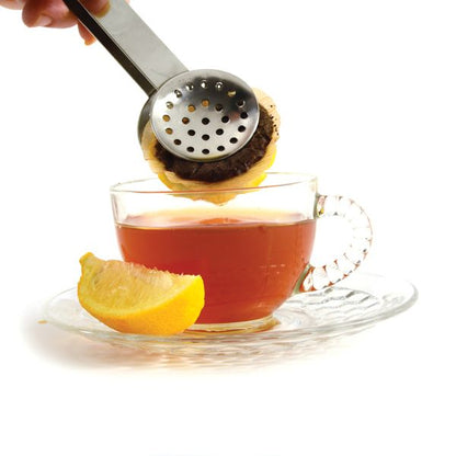 metal tea bag squeezer squeezing a teabag over a clear glass mug of tea and a lemon wedge on a white background