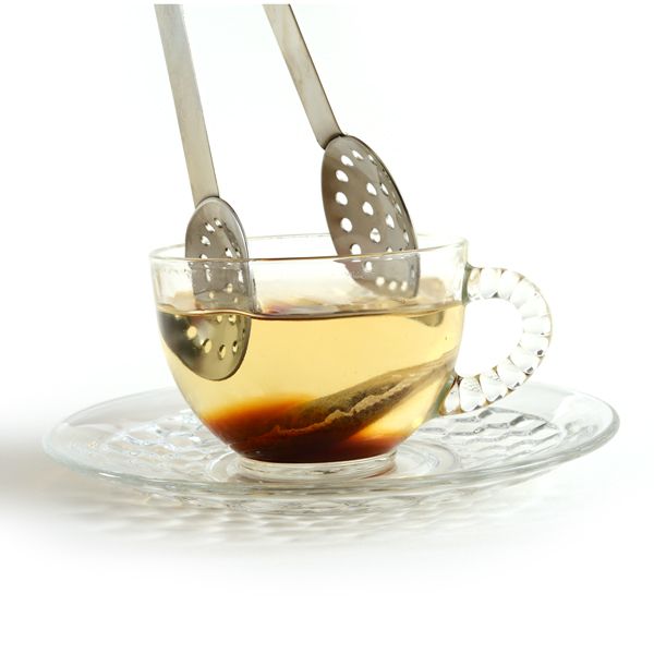 metal tea bag squeezer dipping into a clear glass mug filled with tea and a teabag on a white background.