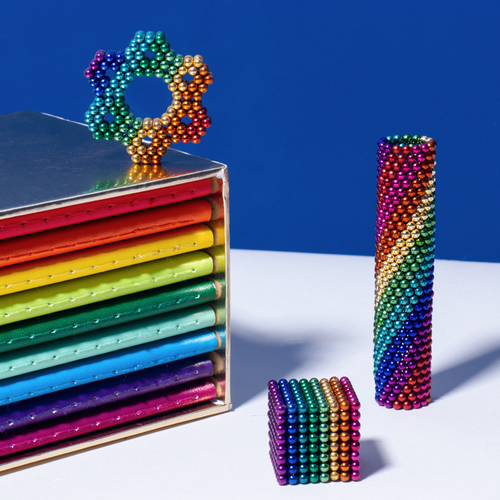 cylinder stack of rainbow colored speks magnet balls, a cube and a flower made from rainbow colored magnetic balls, and a stack of rainbow colored books on a white background.