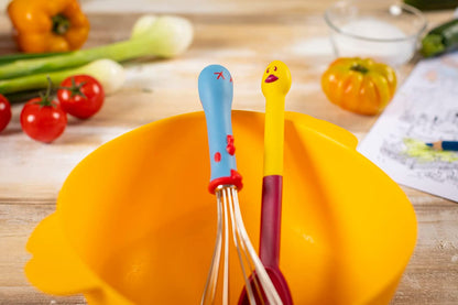 whisk with blue handle and spoon with yellow chicken shaped handle in a yellow bowl on a table with veggies in the background.
