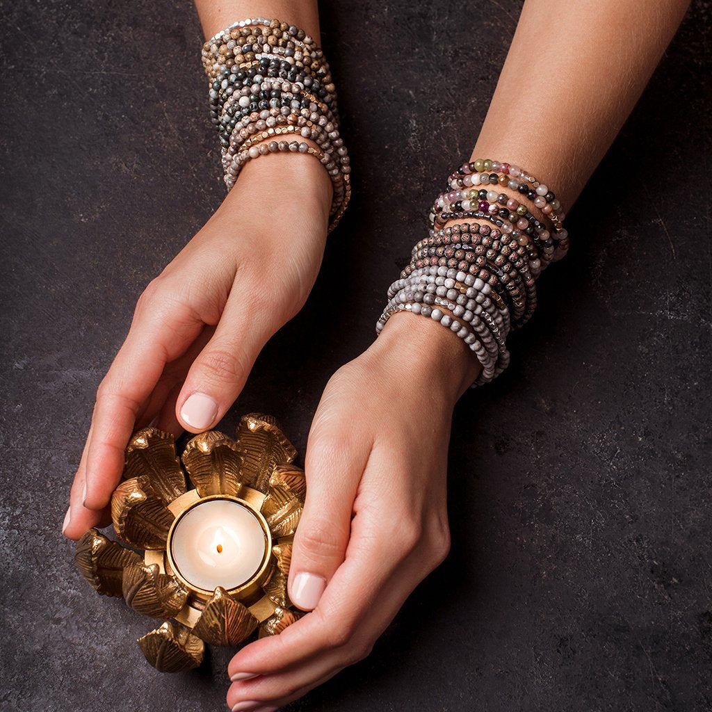 hands holding flower shaped golden votive candle with multiple strands of beaded bracelets on both arms on grey background.