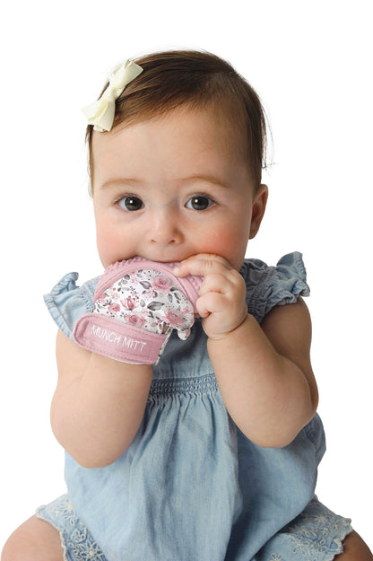 a baby girl with the rosewood munch mitt on one hand and chewing on it against a white background