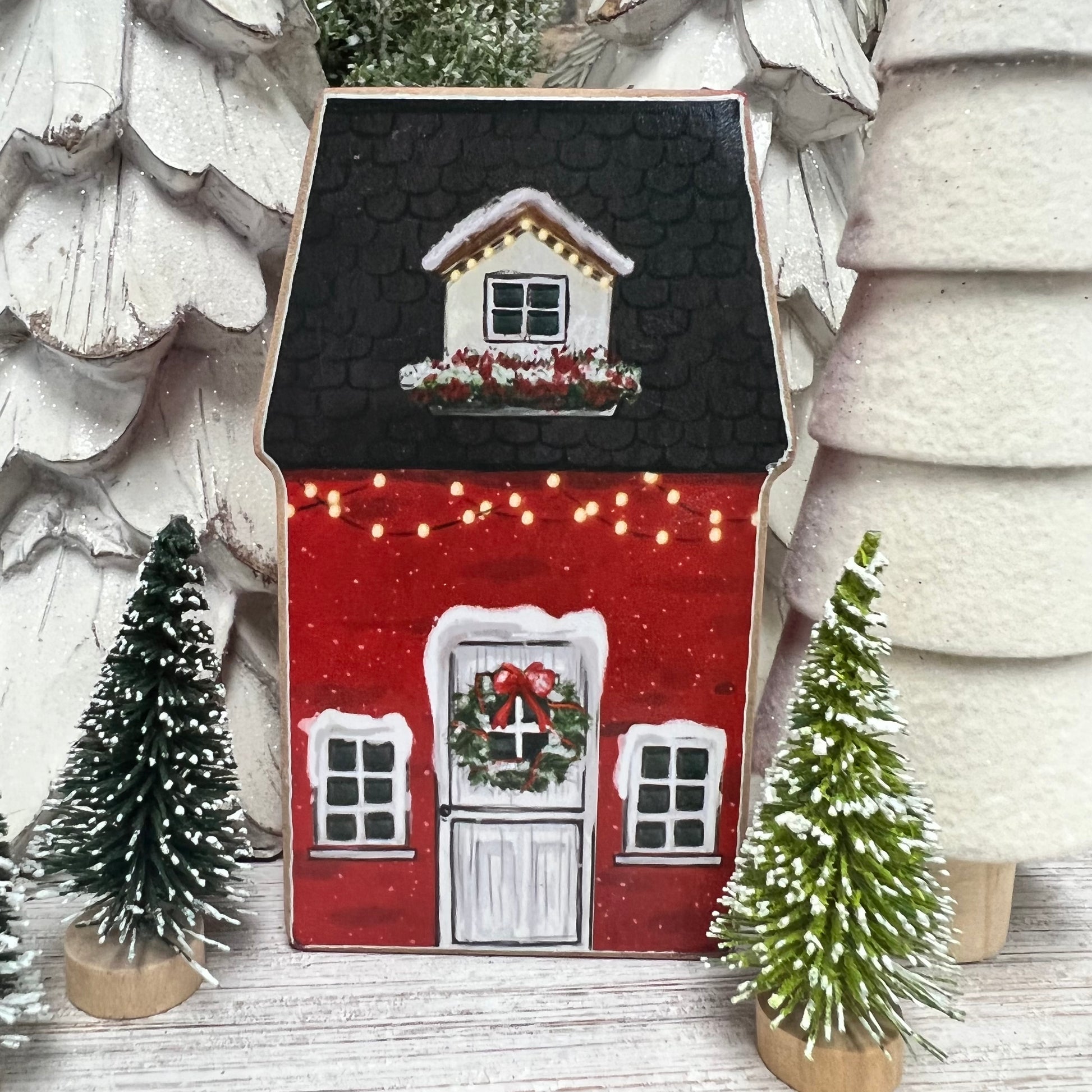 red house painted with white window trim and door with wreath on it and small trees set next to it.