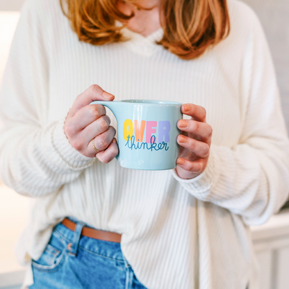 woman wearing white sweater and jeans holding "over thinker" mug.