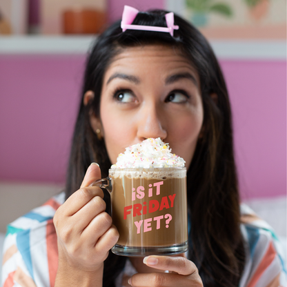 woman with curler in her bangs sipping from "is it friday yet?" glass mug filled with cocoa and whipped cream.