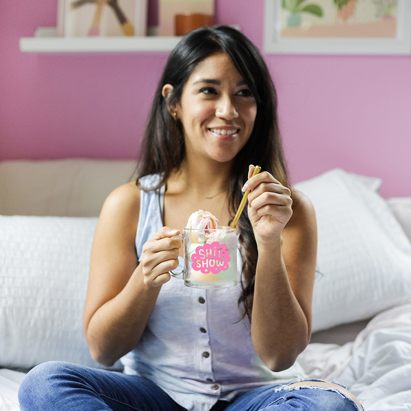 woman sitting on a bed with shit show mug filled with ice cream.