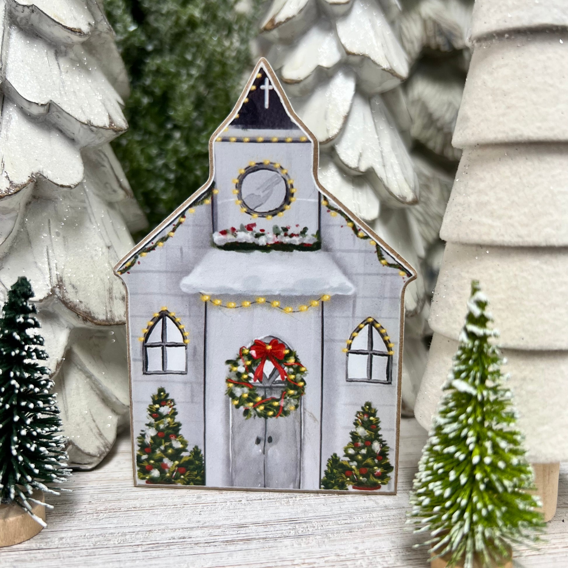 white church painted with wreath on the door, shrubbery decorated with ornaments and lights around the windows with small trees set around it.